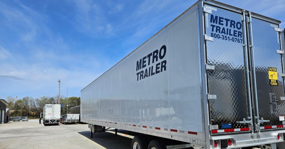 A silver Metro Trailer semi-trailer parked in a paved lot under a blue sky, with another trailer in the distance.