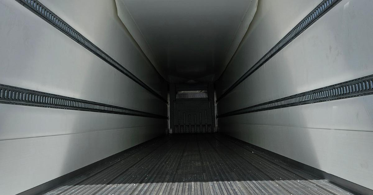 An empty trailer interior with ribbed flooring, white side walls, black rails, and a dim front wall.
