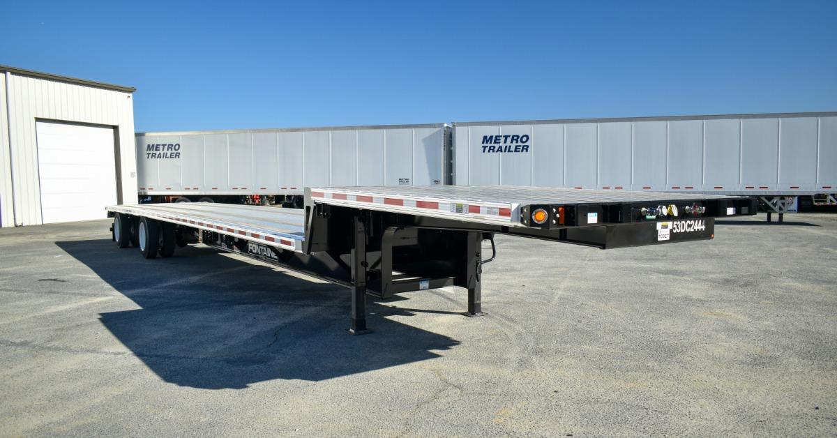 A two-tiered flatbed trailer parked in front of a building and some enclosed trailers. The flatbed is silver with red markings.