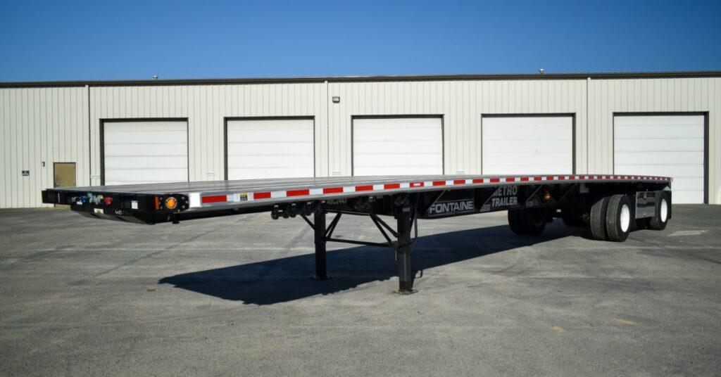 A long flatbed trailer parked in front of some loading bays. The trailer is silver with red dashes along the side.