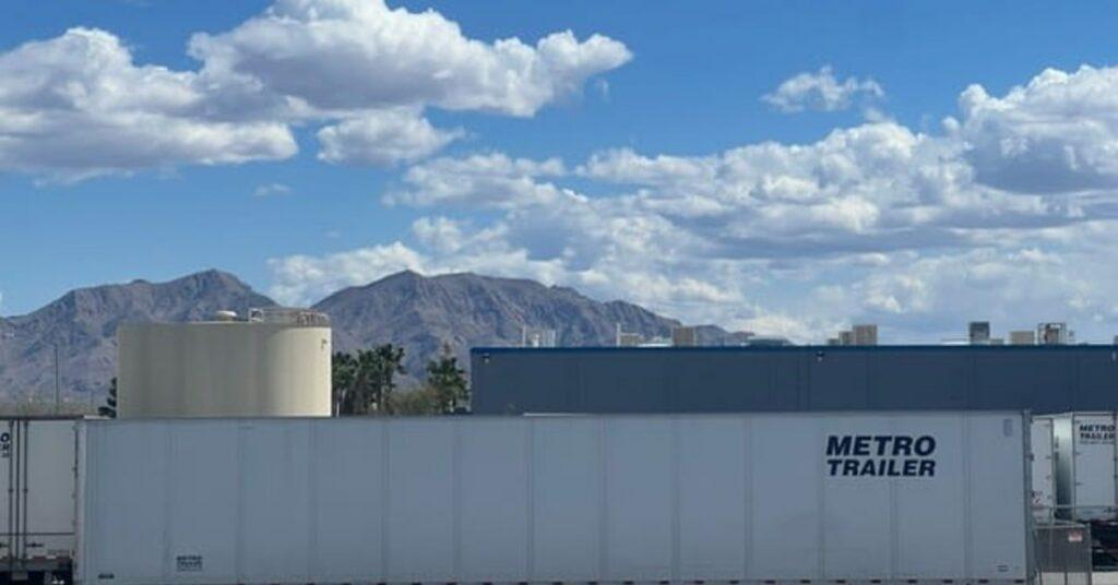 A commercial enclosed trailer parked on a road next to an industrial facility. There are mountains in the background.