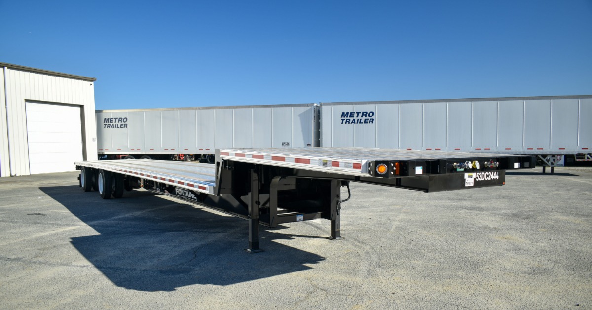 A close-up of a two-tiered flatbed trailer. There are two enclosed trailers parked together behind the flatbed.