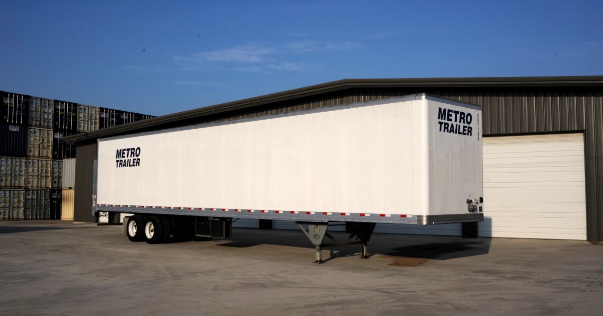 A close-up of an enclosed semi trailer parked next to a building and a large stack of storage containers.