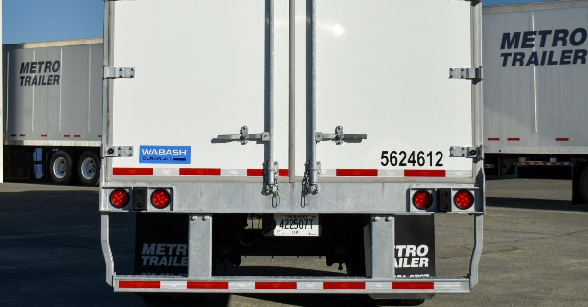 A rear view of a white enclosed trailer with closed cargo doors parked in a trailer lot beneath a clear blue sky.