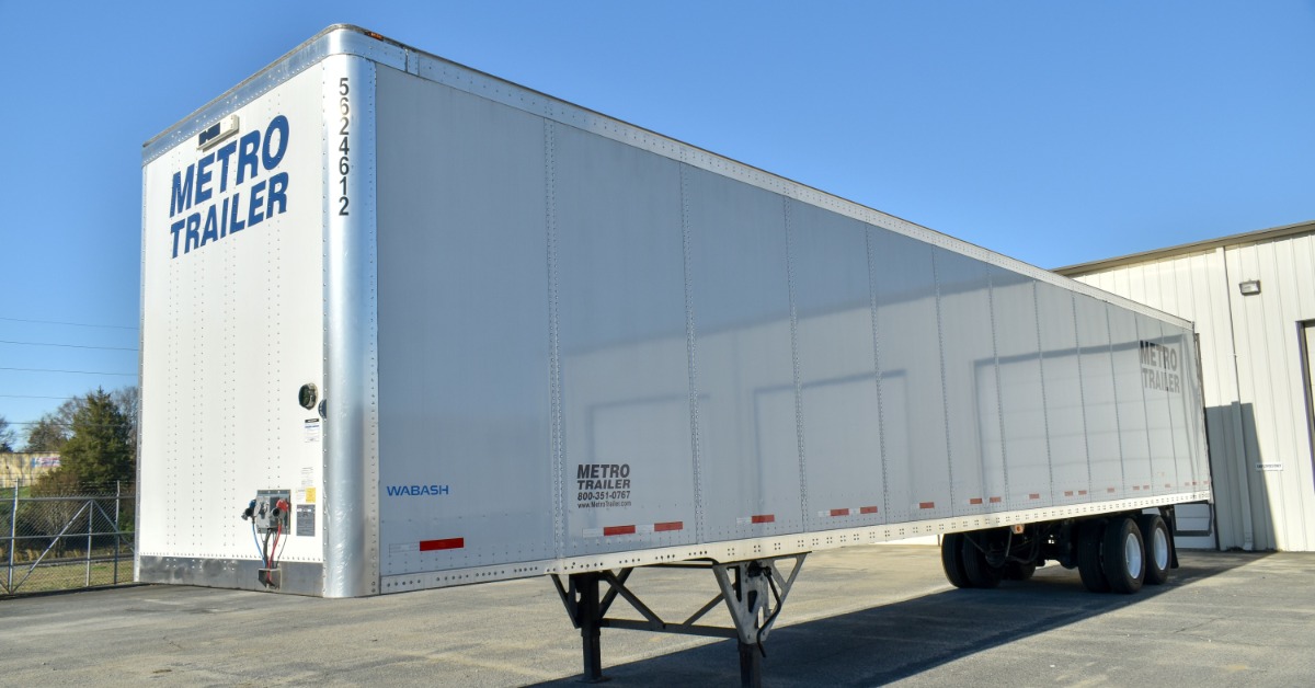 A white semi-trailer with landing gear extended parked outside a warehouse beneath a clear blue sky on a sunny day.