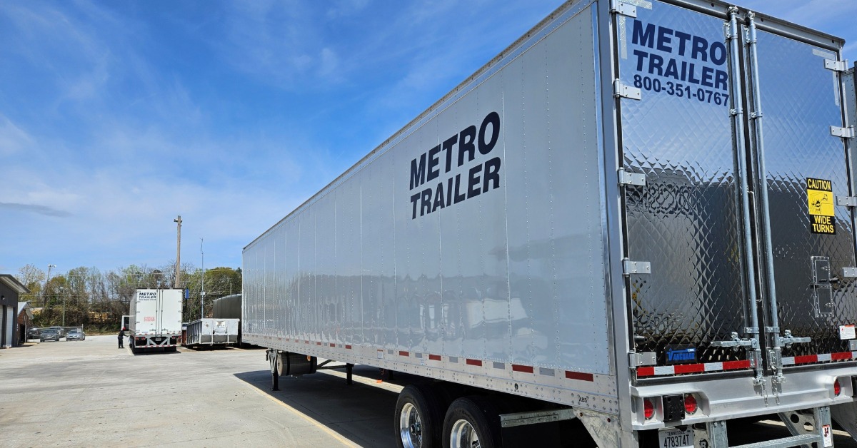 A white enclosed semi-trailer with closed rear doors parked in a commercial lot under a bright blue sky with light clouds.