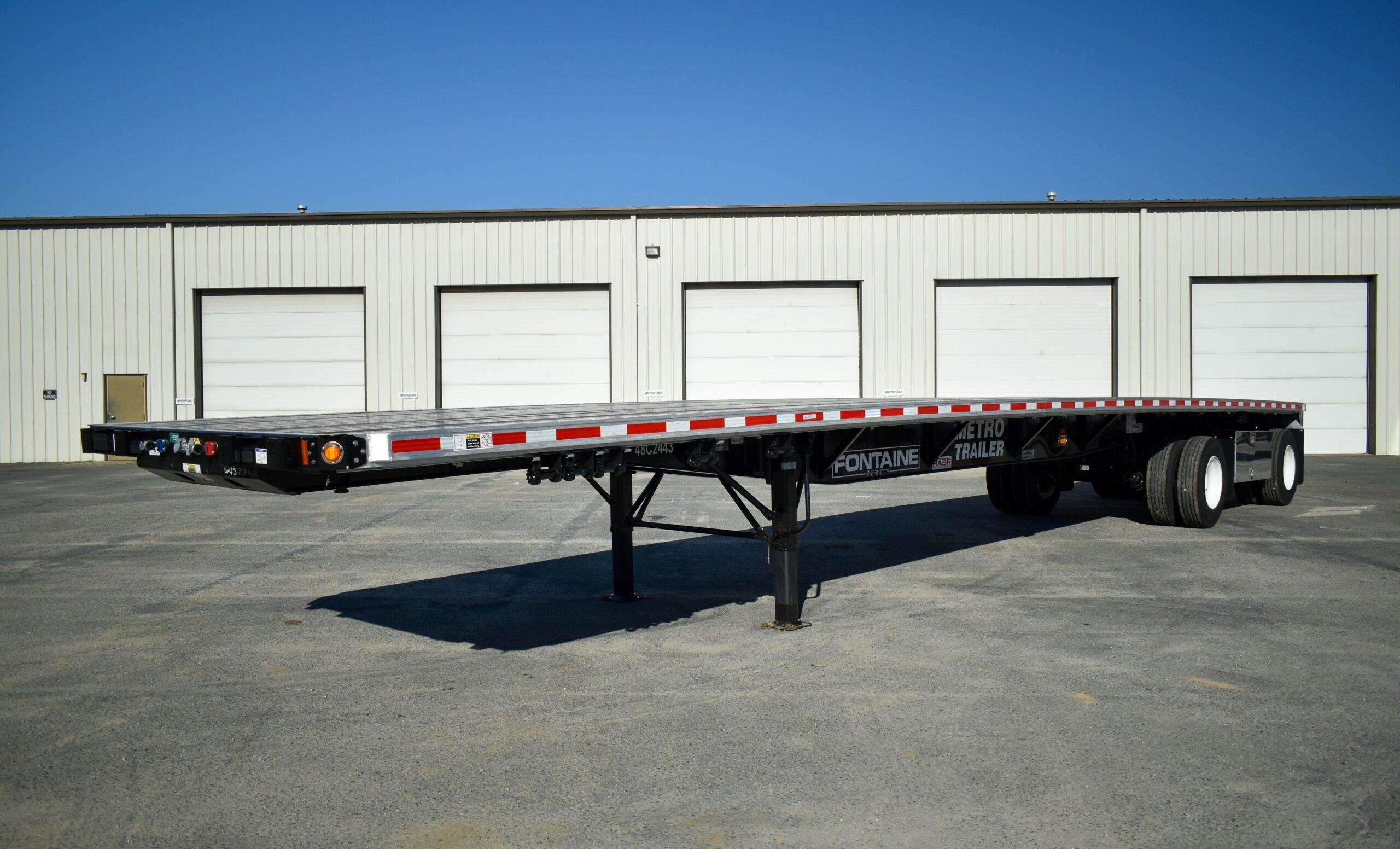 A flatbed semi-trailer parked on pavement in front of a large industrial building with five white garage doors.