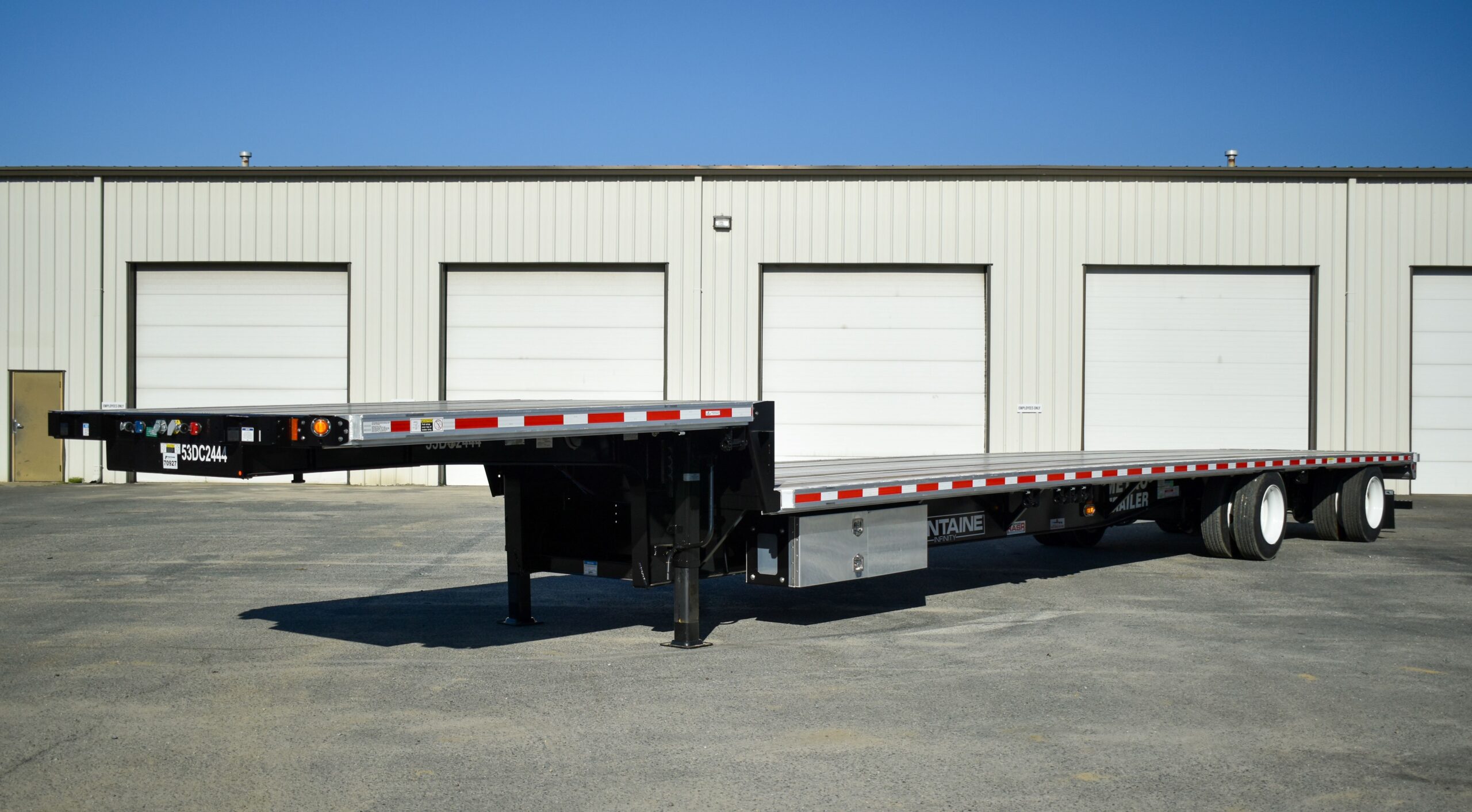 A step deck semi-trailer parked outside an industrial building with several large white garage doors.