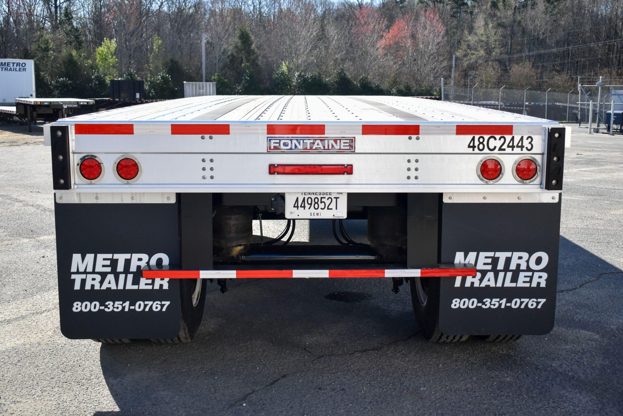 A rear view of a flatbed semi-trailer with red lights, reflective strips, a license plate, and black Metro Trailer mud flaps.