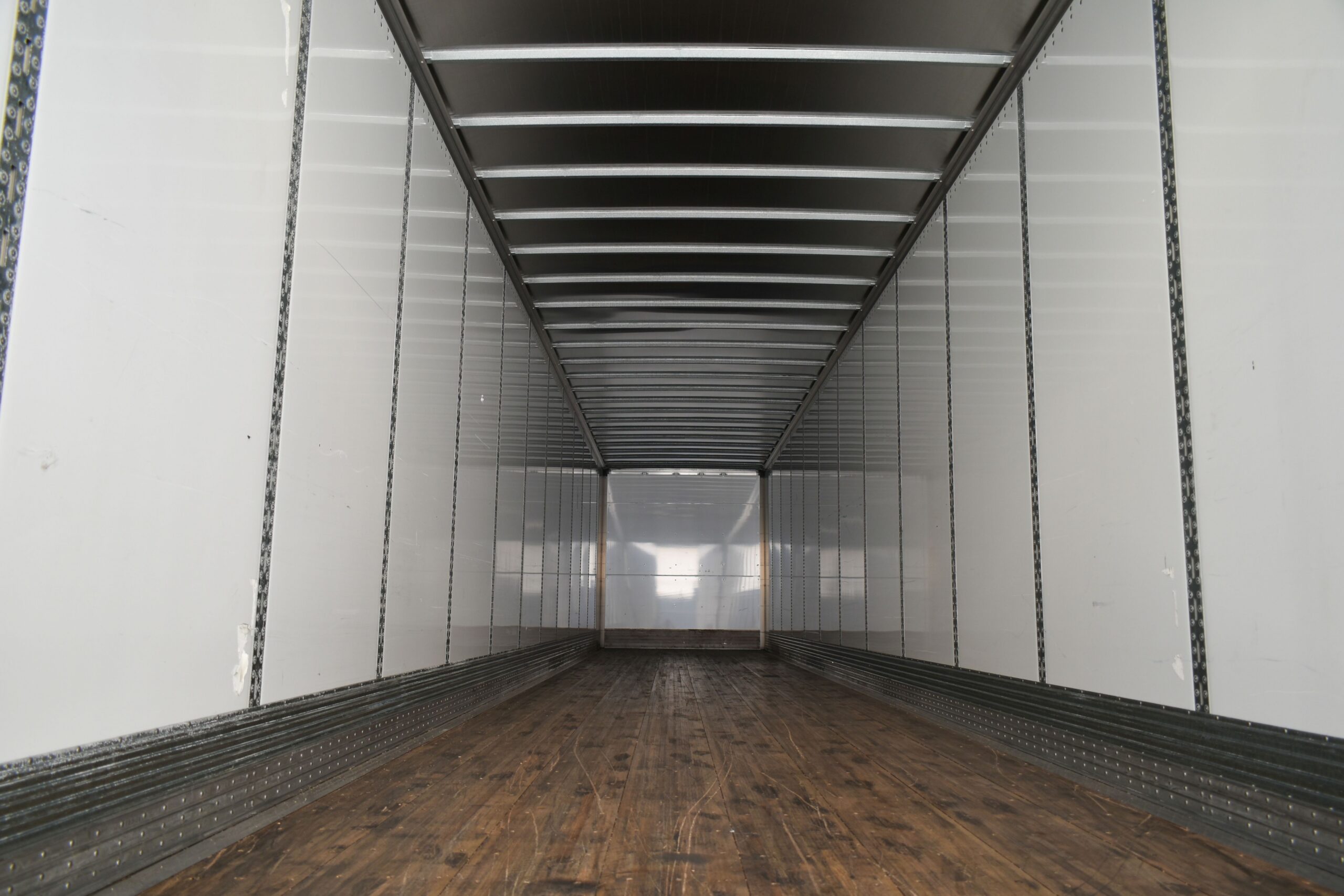 An empty cargo trailer interior with white walls, metal supports, and a long wooden floor extending the length of the trailer.