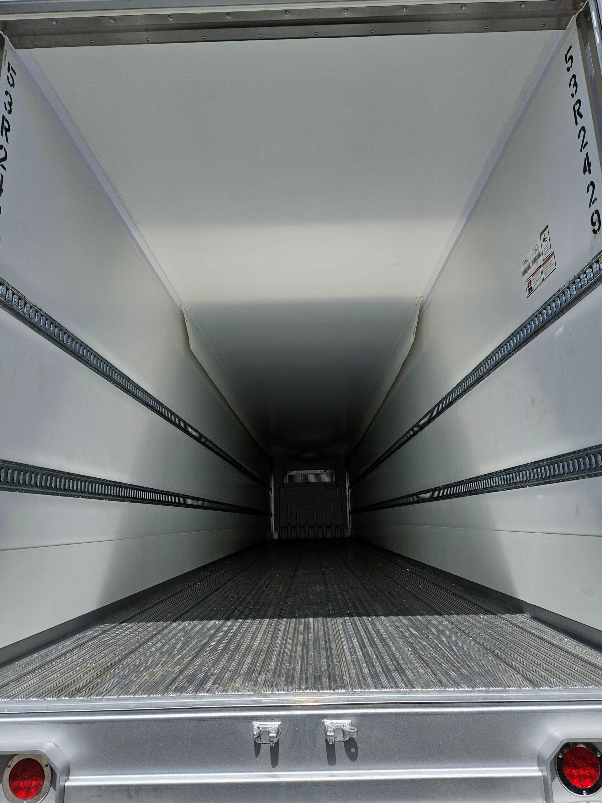 An empty enclosed cargo trailer with white walls, metal cargo rails, and a ribbed floor running along the entire trailer.