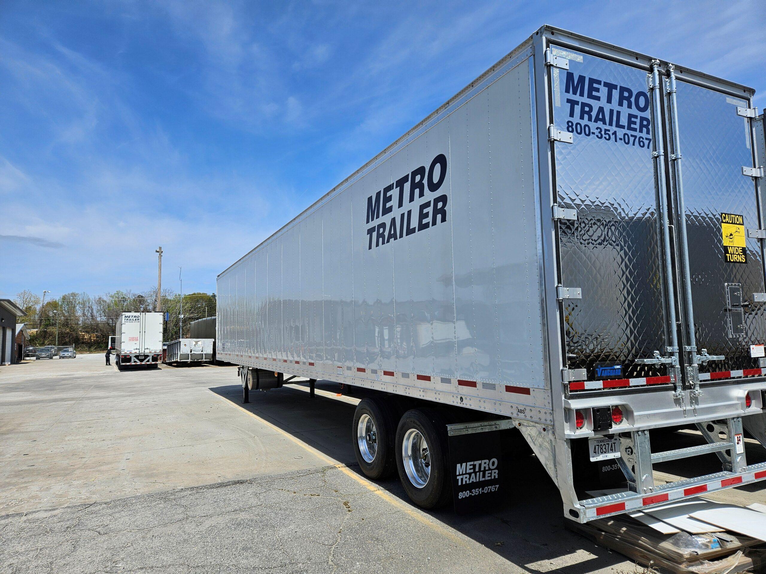 A white and silver Metro Trailer semi-trailer parked in a commercial lot under a bright blue sky with light clouds.