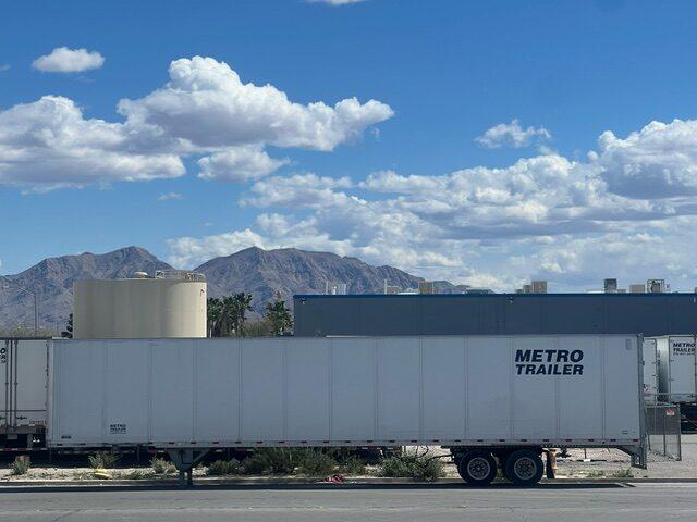 A white Metro Trailer semi-trailer parked outside an industrial facility with mountains and clouds behind it.