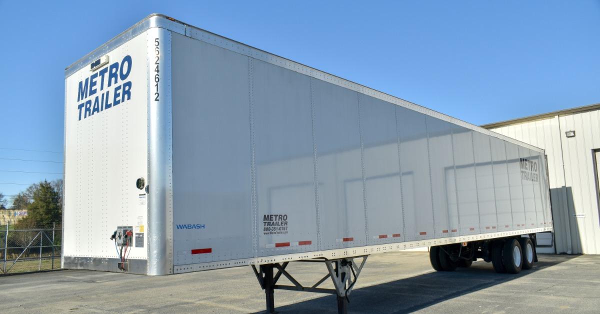 A white Metro Trailer semi-trailer parked on its landing gear outside a warehouse under a clear blue sky.