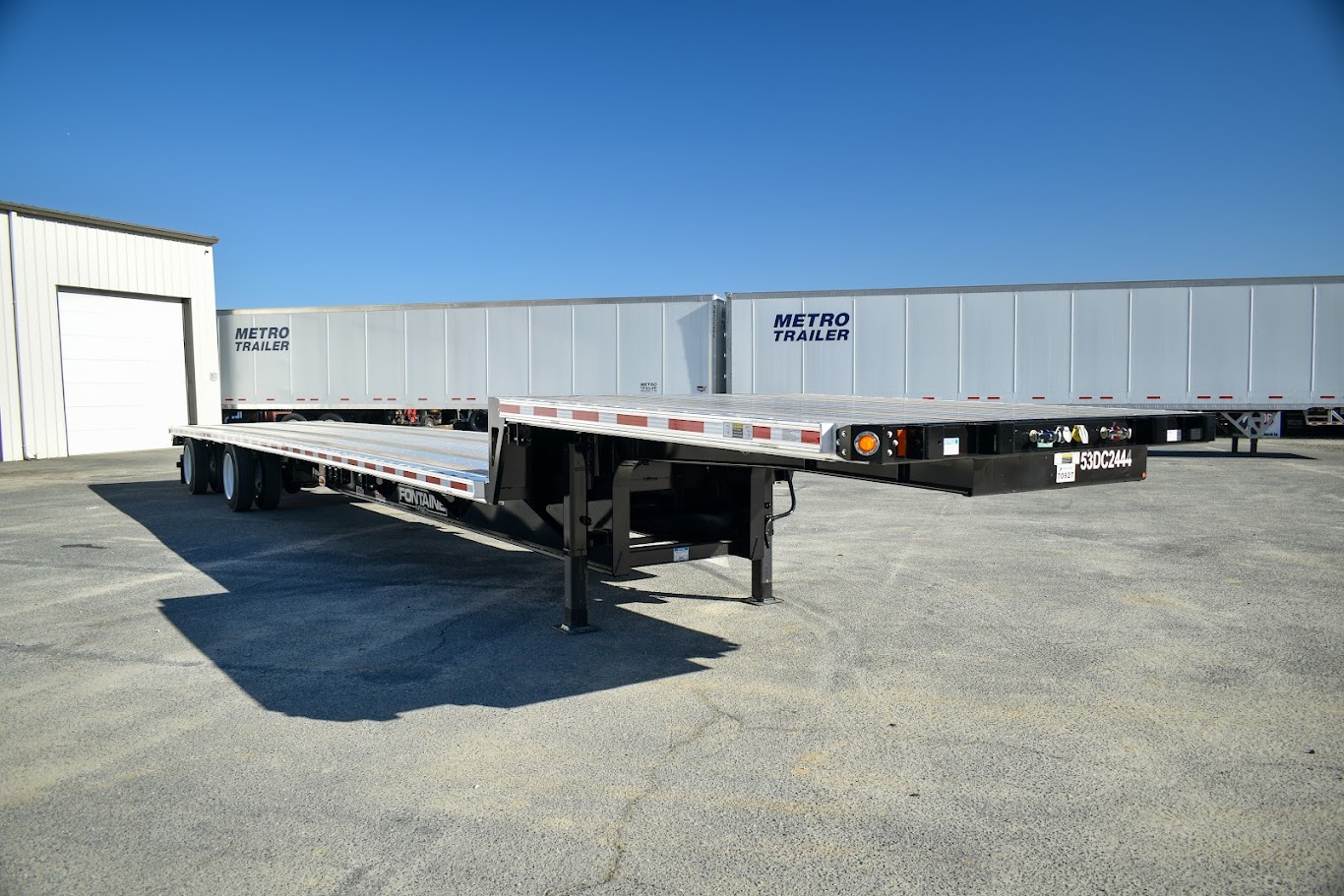 A black and silver flatbed trailer parked on an asphalt lot with two white Metro Trailer semi-trailers behind it.